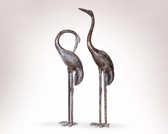 May include: Two bronze crane sculptures. One crane has its head tucked down, while the other stands tall with its neck extended. The sculptures have detailed feather textures and long, textured legs. The background is a gradient of white to light gray.
