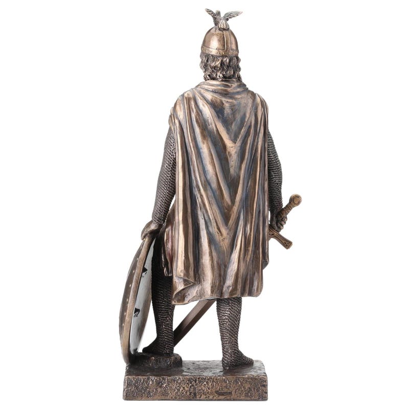May include: Bronze statue of a medieval knight, seen from behind. The knight is clad in a helmet with a bird emblem, chainmail, and a flowing cape. He holds a shield and a sword, standing on a rectangular base. The statue has detailed features.