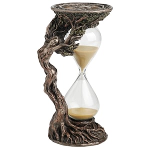 Puede incluir: Un reloj de arena de color bronce con una figura esculpida integrada en una base y una parte superior de tronco de árbol. El cabello y los brazos de la figura se extienden hacia las ramas que sostienen la plataforma superior. El bulbo de vidrio transparente contiene arena fina de color tostado.