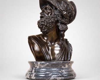 May include: A bronze bust of a bearded man wearing a hat with ornate detailing. The sculpture is mounted on a gray marble base. The man's expression is contemplative, and the bust is a classical art piece.