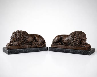 May include: Two bronze lion sculptures in a resting pose, each mounted on a rectangular, dark green marble base. The lions have detailed manes and are a dark brown color. The background is plain white.