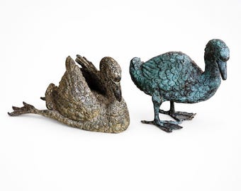May include: Two bronze duck sculptures. One duck is lying down with its wings spread, and the other is standing upright. The duck lying down has a gold and brown finish, while the standing duck has a teal finish.