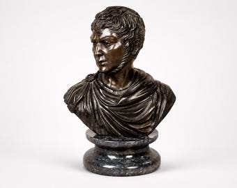 May include: A bronze bust of a man with short, curly hair and a draped garment. The sculpture is mounted on a dark, marble-like pedestal. The man's expression is serious, with a slight furrow of the brow.