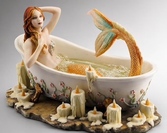 May include: A mermaid figurine in a bathtub, surrounded by candles. The mermaid has red hair and a golden tail. The bathtub is white with floral accents. The candles are white with a yellow glow.