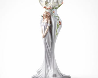 May include: A white ceramic decorative vase with a flared top and a slender, flowing design. The vase features a sculpted figure of a woman in a flowing gown, with floral accents in shades of pink and green. The vase stands approximately 12 inches tall.