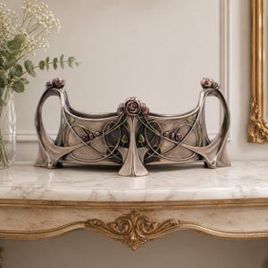 May include: A silver-toned Art Nouveau decorative centrepiece. The piece features a rectangular body with curved handles and is adorned with rose and leaf motifs in shades of pink and green. It sits on a marble-topped table.