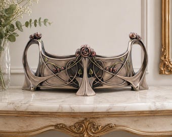 May include: A silver-toned Art Nouveau style decorative centerpiece. The piece features a rectangular body with curved handles and is adorned with rose and leaf motifs in shades of pink and green. It sits on a marble-topped table.