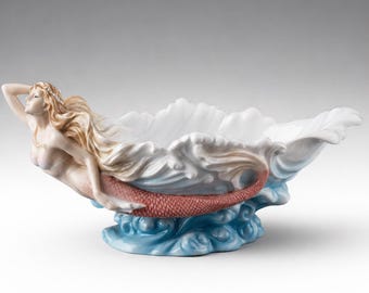 May include: A ceramic mermaid bowl. The bowl is white with a mermaid figure on the side. The mermaid has long blonde hair and a red tail. The bowl is supported by blue waves.
