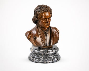 May include: A bronze-toned bust of a man with curly hair and a serious expression. The sculpture is mounted on a black and gray marble base. The bust features detailed facial features and a classic style.