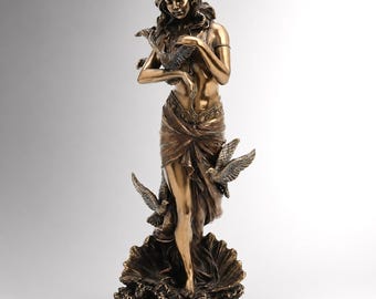 May include: A bronze-colored statue of a woman holding two doves, with two additional doves at her feet. The woman is draped in flowing fabric and stands on a seashell base. The statue is a decorative art piece.