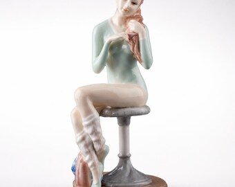 May include: A porcelain figurine of a seated woman with red hair, wearing a light blue leotard and leg warmers. She is seated on a gray stool with a brown base, and is touching her hair. The figurine has a delicate, detailed design.