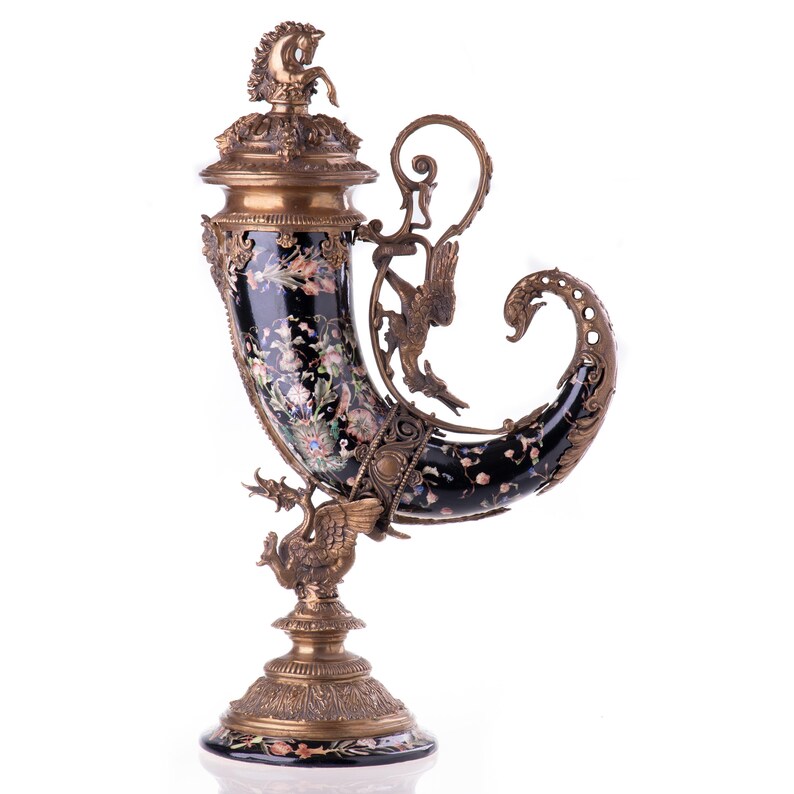 May include: An ornate, antique horn-shaped vase with a black background and floral design. The vase features bronze accents, including a horse figurine on the lid and a swan at the base. The handle has a bird-like design.