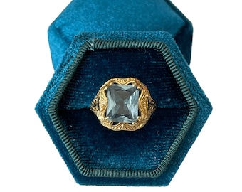 May include: A gold ring with a large, clear, rectangular gemstone set in a decorative, filigree design. The ring is displayed in a teal blue velvet ring box.