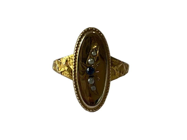 Peut inclure: Une bague en or avec une b&eacute;li&egrave;re ovale sertie d'un saphir bleu et de perles blanches. La bande de la bague est textur&eacute;e avec un motif floral.