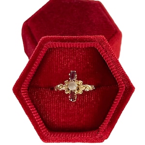 Può includere: Un anello d'oro con una pietra bianca e pietre rosse incastonate a forma di croce. L'anello è in una scatola per anelli in velluto rosso.