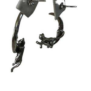 May include: A pair of silver earrings featuring a crescent moon with a face and a cat climbing on it.