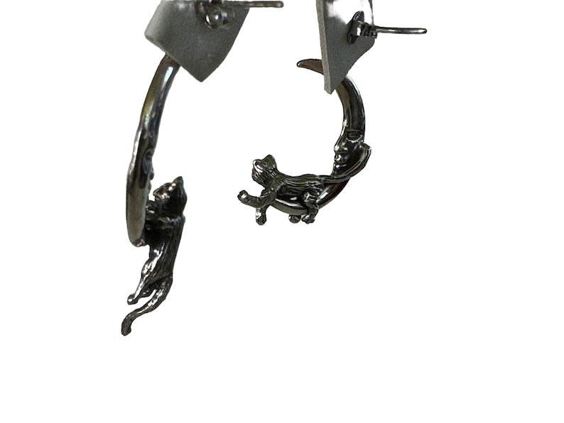 May include: A pair of silver earrings featuring a crescent moon with a face and a cat climbing on it.