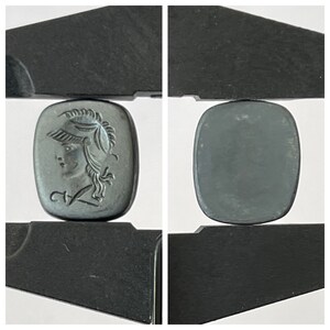 Op de afbeelding: Een zwarte cameo met een gegraveerd beeld van een vrouwenhoofd met een helm. De cameo bevindt zich in een paar zwarte metalen schuifmaten.