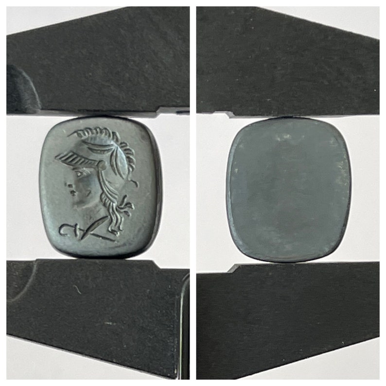 Op de afbeelding: Een zwarte cameo met een gegraveerd beeld van een vrouwenhoofd met een helm. De cameo bevindt zich in een paar zwarte metalen schuifmaten.