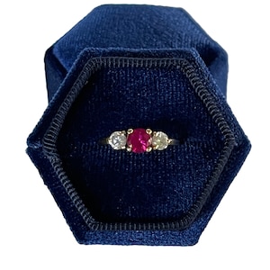 May include: A three-stone ring with a ruby center stone and two smaller diamond side stones. The ring is set in yellow gold and is displayed in a blue velvet ring box.