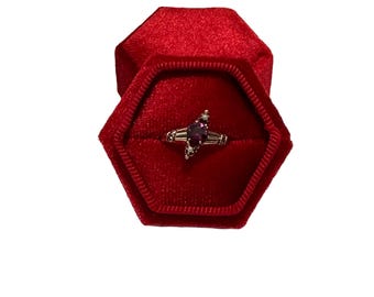 Könnte beinhalten: Ein goldener Ring mit einem lilafarbenen, marquiseförmigen Edelstein und kleinen Akzentsteinen. Der Ring wird in einer offenen, roten sechseckigen Samtbox präsentiert, vor weißem Hintergrund.