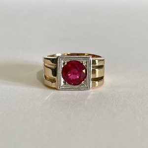 May include: A gold ring with a red gemstone set in a square frame. The ring has a textured band with three raised lines.
