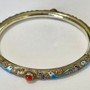 K&ouml;nnte beinhalten: Ein silbernes Armreif-Armband mit blauen und goldenen Emaille-Details und roten Korallen-Akzenten. Das Armband ist mit einem sich wiederholenden Muster aus stilisierten V&ouml;geln und Wellen verziert.