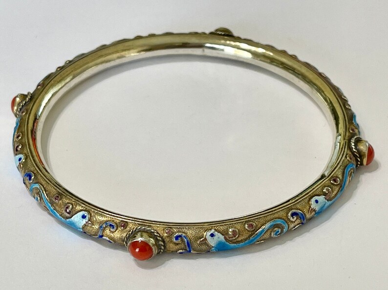 K&ouml;nnte beinhalten: Ein silbernes Armreif-Armband mit blauen und goldenen Emaille-Details und roten Korallen-Akzenten. Das Armband ist mit einem sich wiederholenden Muster aus stilisierten V&ouml;geln und Wellen verziert.