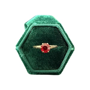 Puede incluir: Un anillo de oro con una gran gema roja redonda se muestra dentro de un joyero hexagonal de terciopelo verde oscuro. La banda del anillo tiene un diseño delicado y texturizado. El joyero está abierto, mostrando el anillo.
