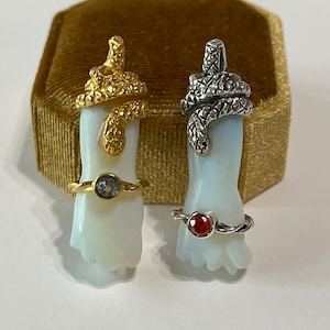 Puede incluir: Dos anillos con bandas en forma de serpiente y piedras preciosas. El anillo de la izquierda tiene una banda de oro con una piedra preciosa transparente. El anillo de la derecha tiene una banda de plata con una piedra preciosa roja.