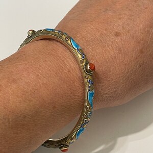 K&ouml;nnte beinhalten: Ein silbernes Armreif-Armband mit blauem und goldenem Emaille und roten Korallenakzenten. Das Armband ist mit einem floralen Design verziert.