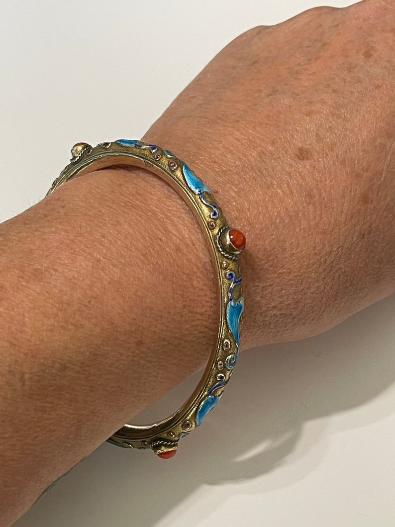 K&ouml;nnte beinhalten: Ein silbernes Armreif-Armband mit blauem und goldenem Emaille und roten Korallenakzenten. Das Armband ist mit einem floralen Design verziert.