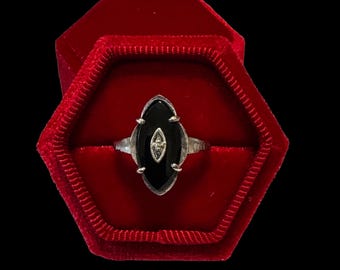 Könnte beinhalten: Ein Silberring mit einem marquiseförmigen schwarzen Onyx in einer Krappenfassung. Ein kleines, längliches silbernes Element mit einem winzigen eingelassenen Stein befindet sich in der Mitte des Onyx. Der Ring liegt in einer sechseckigen roten Samtschatulle.