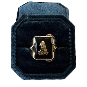Puede incluir: Un anillo de oro con una piedra de ónice negra rectangular engastada en el centro. La piedra está incrustada con una letra A de color dorado en una fuente cursiva decorativa. El aro es de metal dorado pulido y descansa en una caja de terciopelo azul oscuro.