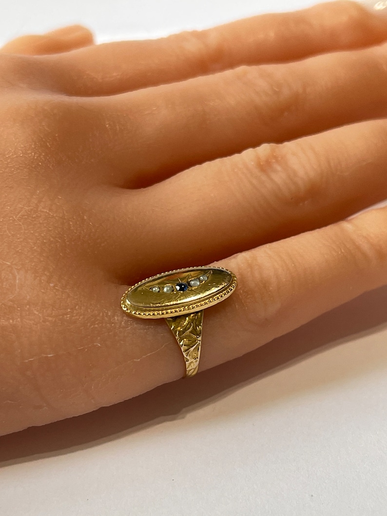 Peut inclure: Une bague en or avec une b&eacute;li&egrave;re ovale sertie de petites perles blanches et d'une seule pierre noire. La bande de la bague est textur&eacute;e.