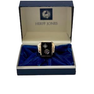 May include: A gold-toned ring with a black onyx center stone, displayed in a blue velvet-lined box. The ring features intricate side detailing and two inset emblems. The box is labeled "HERFF JONES" in white text.
