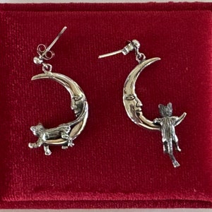 May include: A pair of silver earrings featuring a crescent moon with a face and a cat climbing on it.