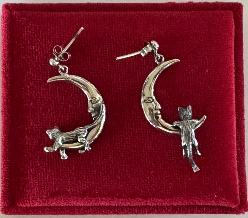 May include: A pair of silver earrings featuring a crescent moon with a face and a cat climbing on it.