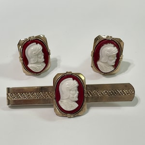 May include: A set of gold-toned cufflinks and a tie bar featuring red and white cameo portraits of a man in a helmet.