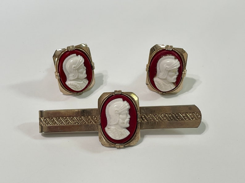 May include: A set of gold-toned cufflinks and a tie bar featuring red and white cameo portraits of a man in a helmet.