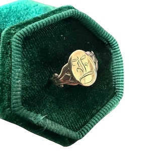 May include: A gold signet ring with an engraved letter "F" on an oval face, presented in a dark green velvet ring box. The ring features decorative detailing on the band and is set against a white backdrop.
