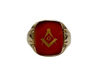 May include: A gold-toned ring with a rectangular, red stone. The stone features a gold-colored Masonic symbol, including a compass, square, and the letter "G". The ring has a detailed band design.