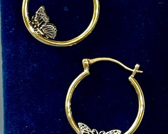 May include: A pair of gold hoop earrings with a butterfly design. The butterflies are silver-colored and have intricate cut-out details. The earrings are displayed on a dark blue velvet surface.