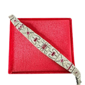 May include: A silver-toned bracelet featuring a geometric metalwork pattern set with rectangular, deep pink-red gemstones. The design incorporates intricate filigree-style detailing and small clear stones throughout the metal links.