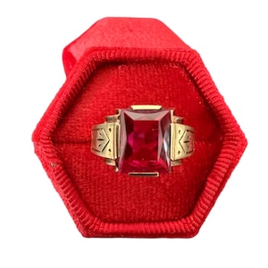 May include: A gold-toned ring with a large, rectangular, red gemstone. The ring is set in a red velvet box. The ring has decorative side accents. The gemstone is the focal point of the ring.