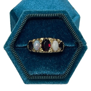 May include: A gold ring featuring three oval-shaped gemstones. The central stone is a deep red garnet, with two white opals on either side. The ring is set in a gold band and is presented in a teal velvet ring box.