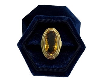May include: A gold ring with an oval, amber-colored gemstone, set in a gold band. The ring is displayed in a dark blue, hexagonal velvet ring box. The ring's design is simple and elegant.