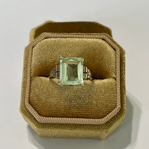 May include: A green gemstone ring with a square-shaped stone set in a gold band. The ring is in a small, velvet-lined box.