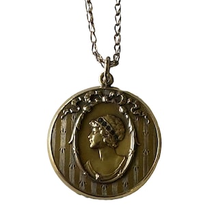 May include: A gold locket necklace with a detailed design featuring a woman's profile. The locket has a chain link design and a decorative border.