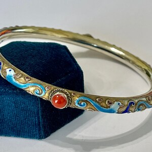K&ouml;nnte beinhalten: Ein silbernes Armreif-Armband mit blauen und wei&szlig;en Emaildetails und roten Korallenakzenten. Das Armband ist mit einem wirbelnden Muster und V&ouml;geln verziert.